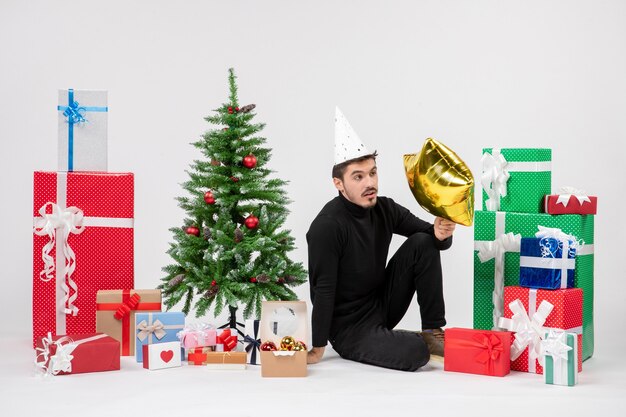 Vue de face du jeune homme assis autour de cadeaux et tenant une étoile d'or sur un mur blanc