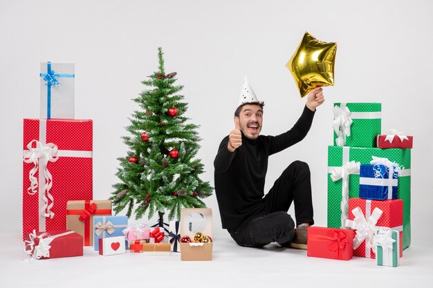 Vue de face du jeune homme assis autour de cadeaux et tenant une étoile d'or sur un mur blanc