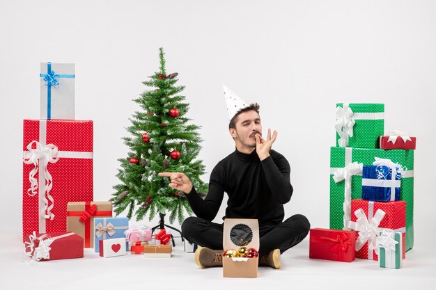 Vue de face du jeune homme assis autour des cadeaux de vacances sur le mur blanc