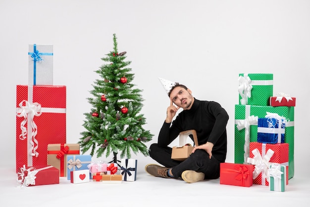 Vue de face du jeune homme assis autour des cadeaux de vacances sur le mur blanc