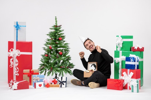 Vue de face du jeune homme assis autour des cadeaux de vacances sur le mur blanc