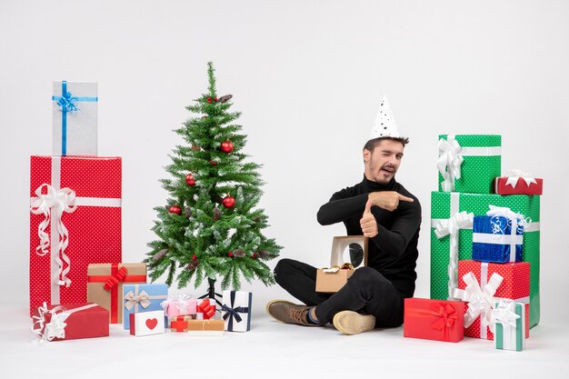 Vue de face du jeune homme assis autour des cadeaux de vacances sur le mur blanc