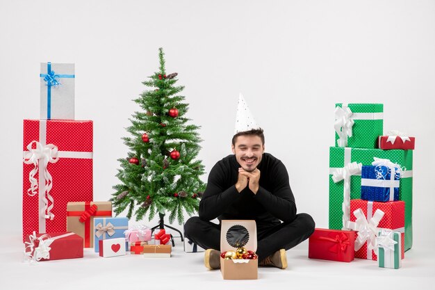 Vue de face du jeune homme assis autour des cadeaux de vacances sur le mur blanc