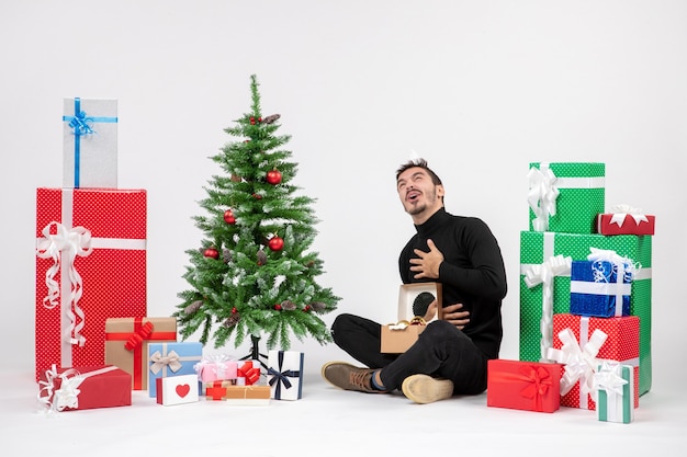 Vue de face du jeune homme assis autour des cadeaux de vacances sur le mur blanc