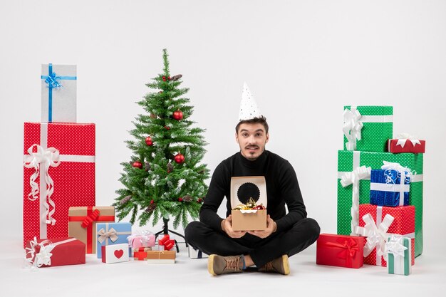 Vue de face du jeune homme assis autour des cadeaux de vacances tenant des jouets d'arbre sur un mur blanc