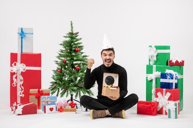 Vue de face du jeune homme assis autour des cadeaux de vacances tenant des jouets d'arbre sur un mur blanc