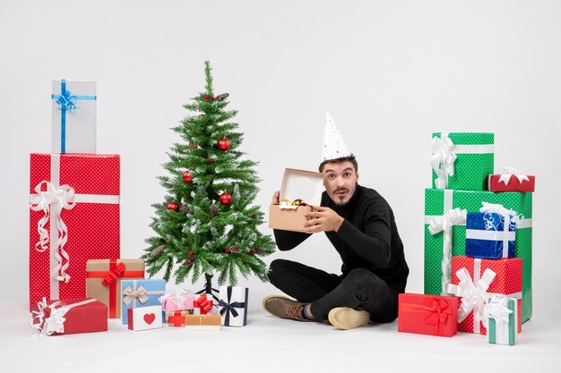 Vue de face du jeune homme assis autour de vacances cadeaux holding package avec jouets sur mur blanc