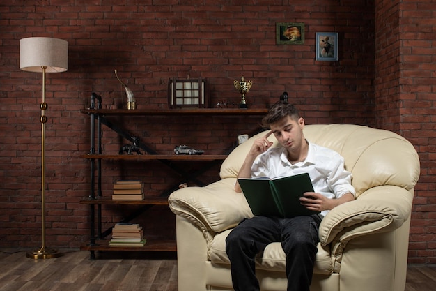 Vue de face du jeune homme assis sur le canapé livre de lecture à l'intérieur de la chambre travail travail soirée
