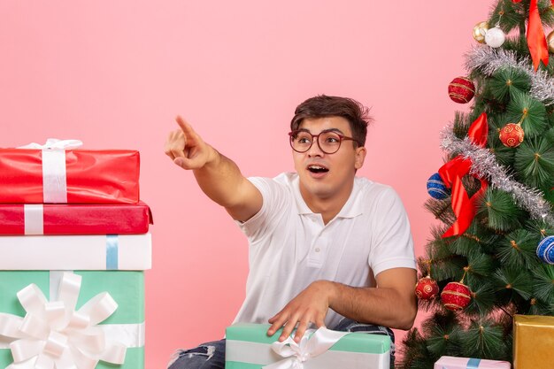 Vue de face du jeune homme autour de cadeaux et arbre de Noël sur le mur rose