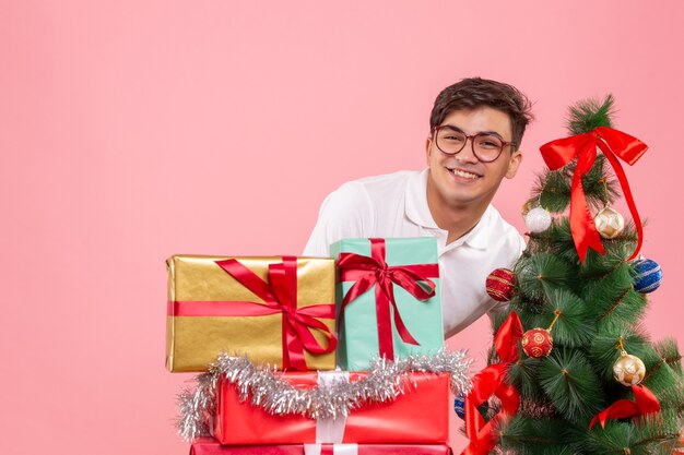 Vue de face du jeune homme autour des cadeaux de Noël et arbre de vacances sur mur rose