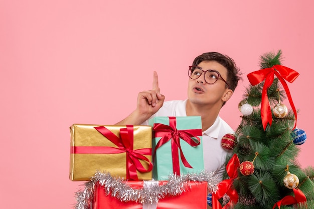 Vue de face du jeune homme autour des cadeaux de Noël et arbre de vacances sur mur rose