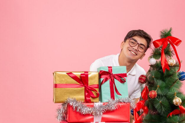 Vue de face du jeune homme autour des cadeaux de Noël et arbre de vacances sur mur rose