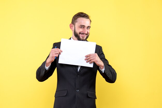 Vue de face du jeune homme beau jeune homme souriant et tenant un papier blanc vierge avec un stylo sur jaune