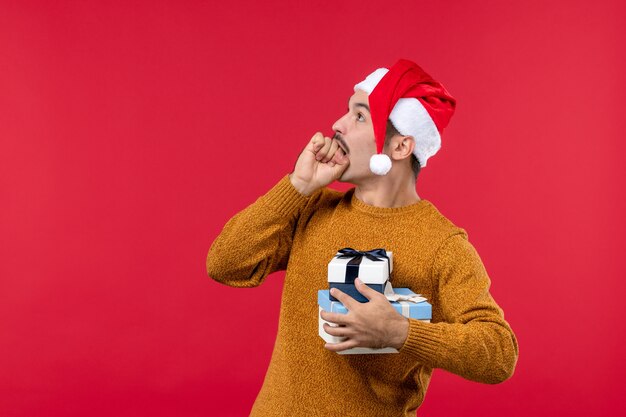 Vue de face du jeune homme avec des cadeaux de nouvel an sur le mur rouge