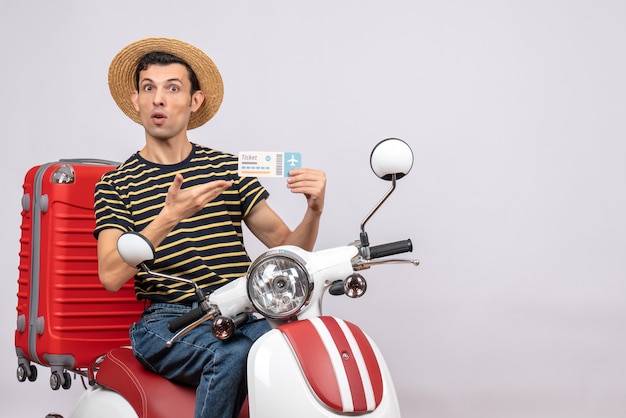 Vue de face du jeune homme avec chapeau de paille sur cyclomoteur pointant sur billet d'avion