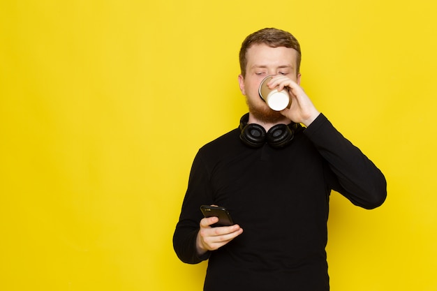 Vue de face du jeune homme en chemise noire à l'aide de téléphone et de boire du café