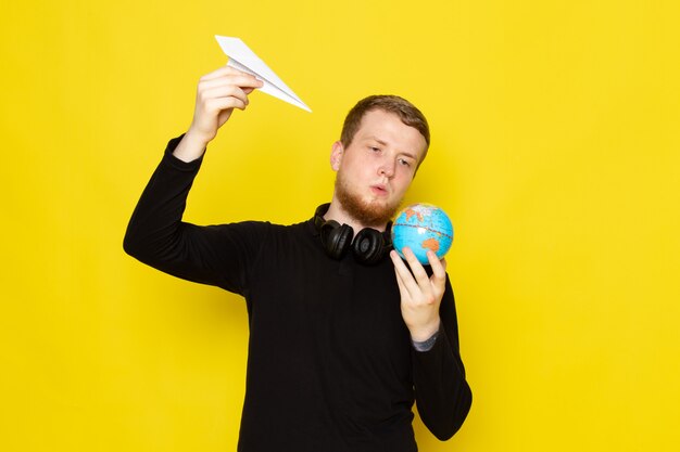 Vue de face du jeune homme en chemise noire tenant un avion en papier et une petite forme de globe