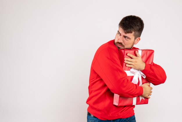 Vue de face du jeune homme en chemise rouge protégeant le cadeau de Noël sur le mur blanc