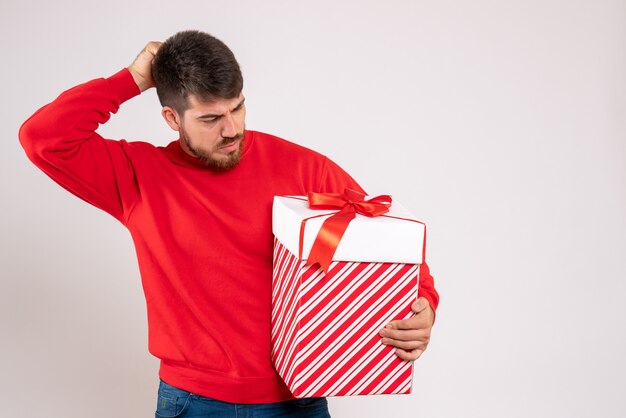 Vue de face du jeune homme en chemise rouge tenant le cadeau de Noël dans la boîte sur le mur blanc