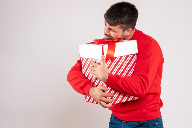 Vue de face du jeune homme en chemise rouge tenant le cadeau de Noël dans la boîte sur le mur blanc