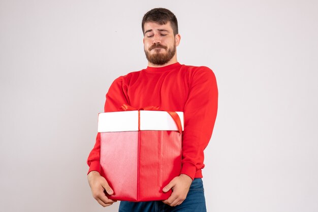 Vue de face du jeune homme en chemise rouge tenant un cadeau de Noël dans une boîte sur un mur blanc