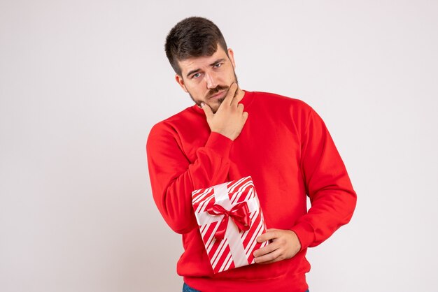 Vue de face du jeune homme en chemise rouge tenant le cadeau de Noël sur le mur blanc