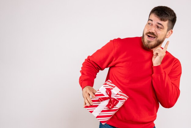 Vue de face du jeune homme en chemise rouge tenant le cadeau de Noël sur le mur blanc