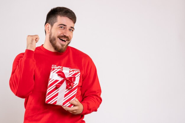 Vue de face du jeune homme en chemise rouge tenant le cadeau de Noël sur le mur blanc