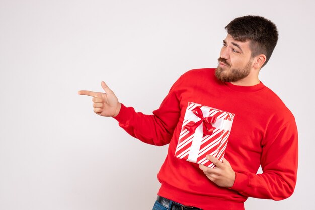 Vue de face du jeune homme en chemise rouge tenant le cadeau de Noël sur le mur blanc