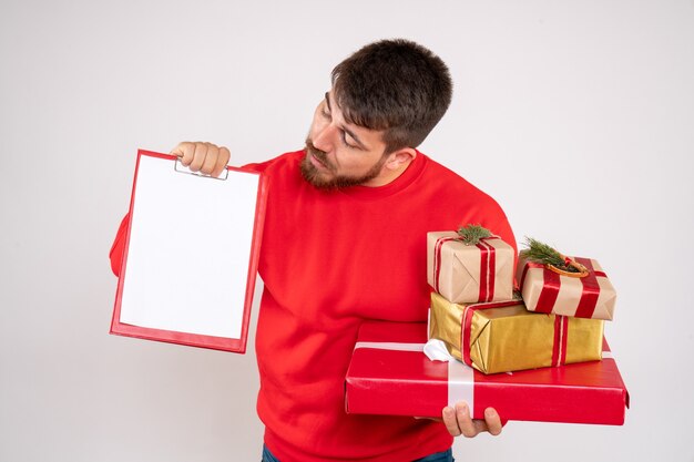 Vue de face du jeune homme en chemise rouge tenant des cadeaux de Noël et note de fichier sur mur blanc