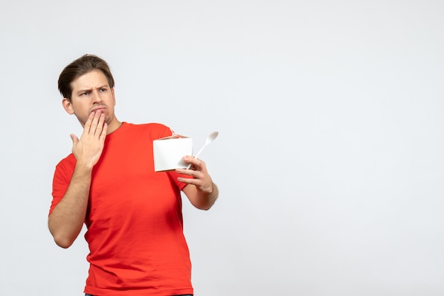 Vue de face du jeune homme choqué en chemisier rouge tenant une boîte de papier et une cuillère sur fond blanc