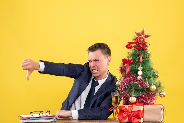 Vue de face du jeune homme en colère faisant signe du pouce vers le bas assis à la table près de l'arbre de Noël et des cadeaux sur le mur jaune