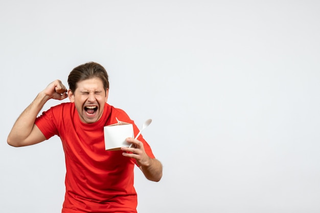 Vue de face du jeune homme émotionnel heureux en chemisier rouge tenant une boîte de papier et une cuillère sur fond blanc