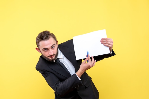 Vue de face du jeune homme jeune homme d'affaires pointant avec un stylo sur un papier vierge de côté vers la gauche sur jaune