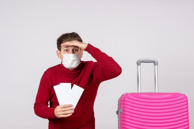 Vue de face du jeune homme en masque tenant des billets d'avion sur un mur blanc