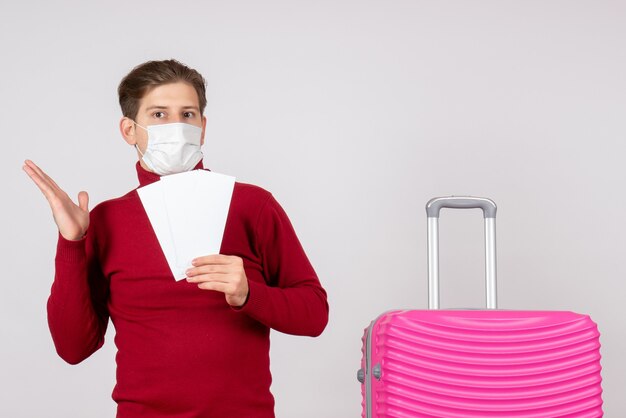 Vue de face du jeune homme en masque tenant des billets d'avion sur un mur blanc