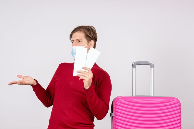 Vue de face du jeune homme en masque tenant des billets d'avion sur un mur blanc