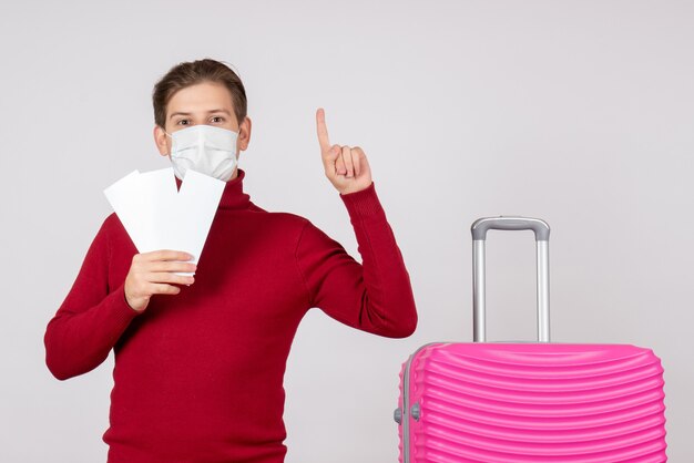 Vue de face du jeune homme en masque tenant des billets d'avion sur un mur blanc