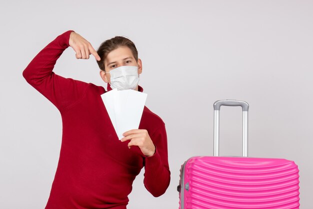 Vue de face du jeune homme en masque tenant des billets d'avion sur un mur blanc