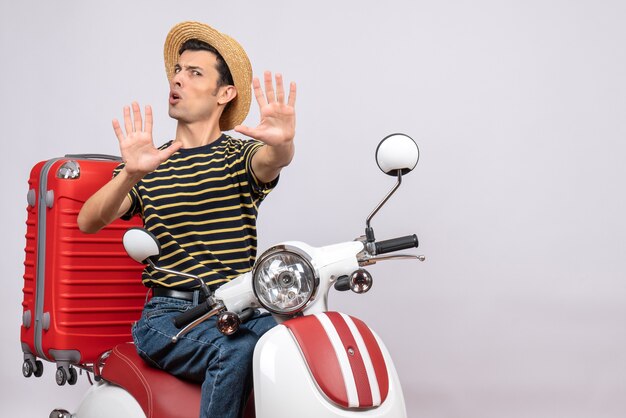 Vue de face du jeune homme mécontent avec chapeau de paille sur cyclomoteur faisant panneau d'arrêt