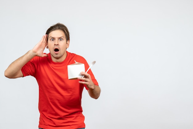 Vue de face du jeune homme perplexe en chemisier rouge tenant une boîte de papier et une cuillère sur fond blanc