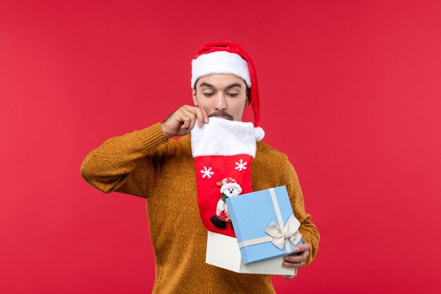 Vue de face du jeune homme prenant la chaussette de Noël de la boîte sur le mur rouge