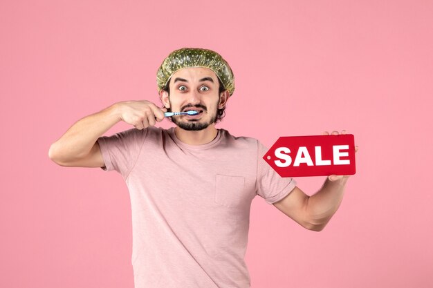 vue de face du jeune homme se nettoyant les dents et tenant une bannière de vente sur un mur rose