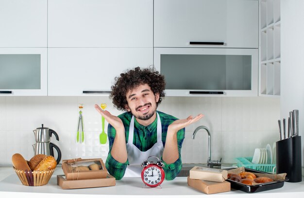 Vue de face du jeune homme souriant debout derrière l'horloge de table diverses pâtisseries dessus dans la cuisine blanche