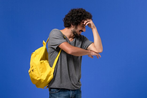 Vue de face du jeune homme en t-shirt gris portant un sac à dos jaune juste en posant et en riant sur le mur bleu