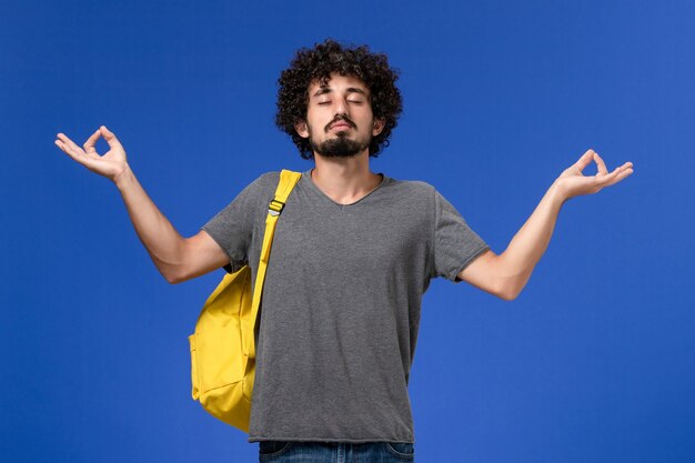 Vue de face du jeune homme en t-shirt gris portant un sac à dos jaune méditant sur le mur bleu