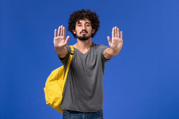 Vue de face du jeune homme en t-shirt gris portant un sac à dos jaune montrant ses paumes sur mur bleu