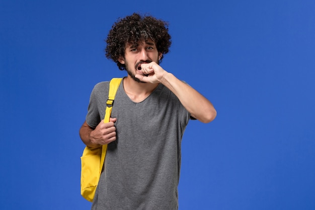Vue de face du jeune homme en t-shirt gris portant un sac à dos jaune sur le mur bleu