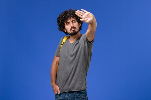 Vue de face du jeune homme en t-shirt gris portant un sac à dos jaune sur le mur bleu