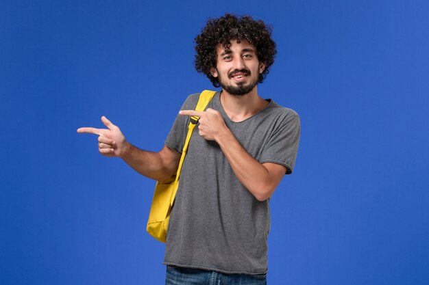 Vue de face du jeune homme en t-shirt gris portant un sac à dos jaune souriant juste sur le mur bleu